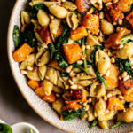 a close up shot of the pasta in a bowl tossed with butternut squash and spinach garnished with vegan parmesan cheese and toasted pine nuts