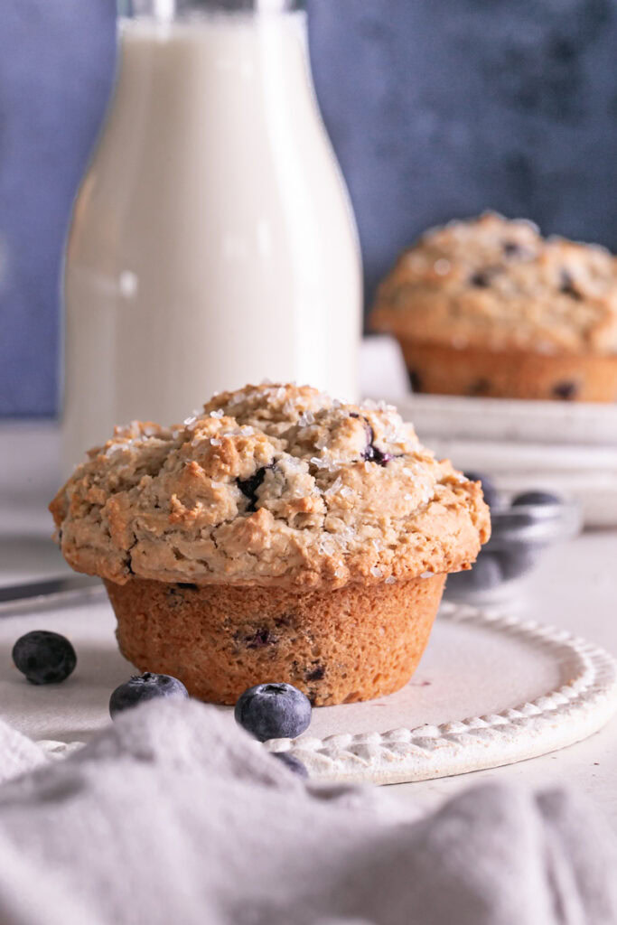 one blueberry muffin on a plate with a jar of almond milk in the background and another muffin on a stack of plates with fresh blueberries around it