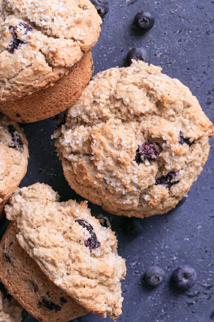 close up of blueberry muffins on a blueberry colored backdrop with fresh blueberries around the muffins