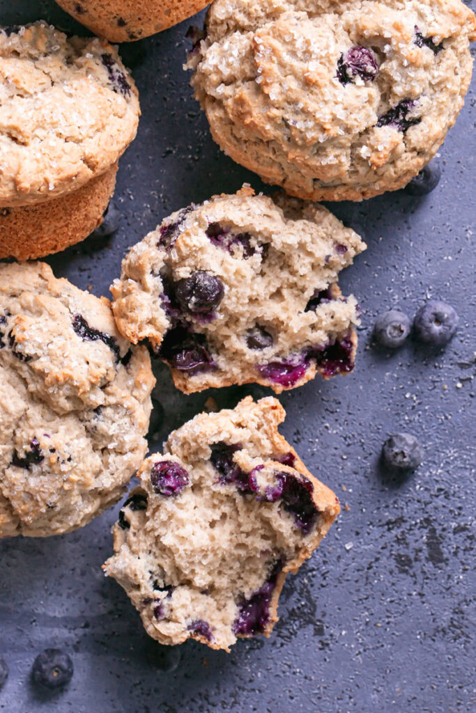 blueberry muffins on a blueberry colored backdrop with fresh blueberries around the muffins and one of the muffins cut in half to reveal the soft inside