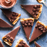slices of Twix tart piped with chocolate mousse on a blue backdrop with a bowl of chocolate mousse and a bowl of vegan caramel