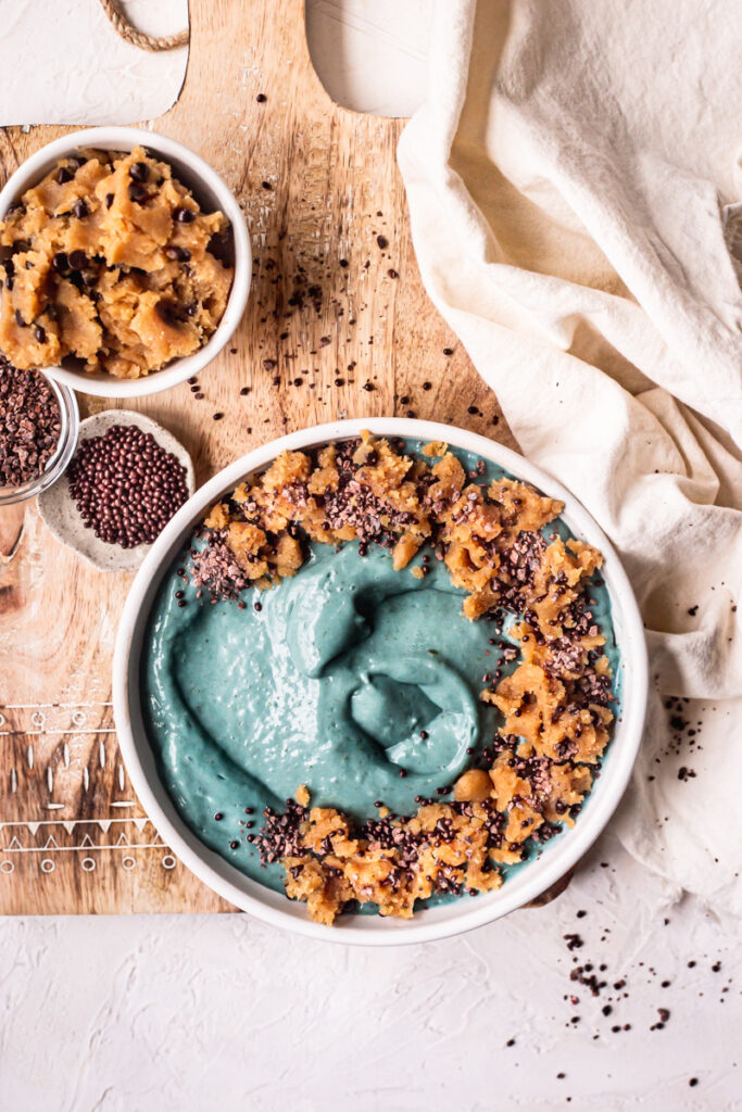 a blue spirulina smoothie bowl topped with almond flour cookie dough, chocolate covered chia seeds on top of a wooden cutting board with a bowl of cookie dough and cacao nibs beside it.