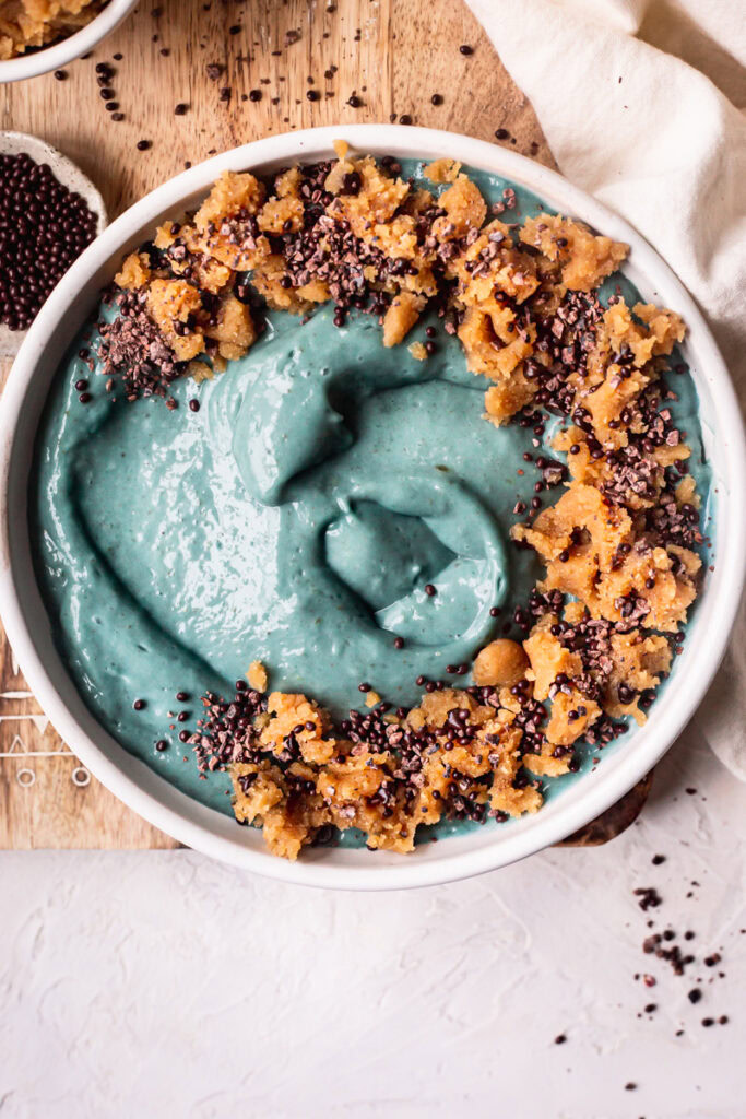 a blue spirulina smoothie bowl topped with almond flour cookie dough, chocolate covered chia seeds on top of a wooden cutting board with a bowl of cookie dough and cacao nibs beside it.