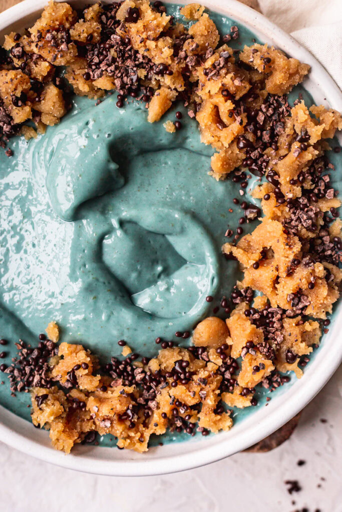 a close up of a blue spirulina smoothie bowl topped with almond flour cookie dough, chocolate covered chia seeds.