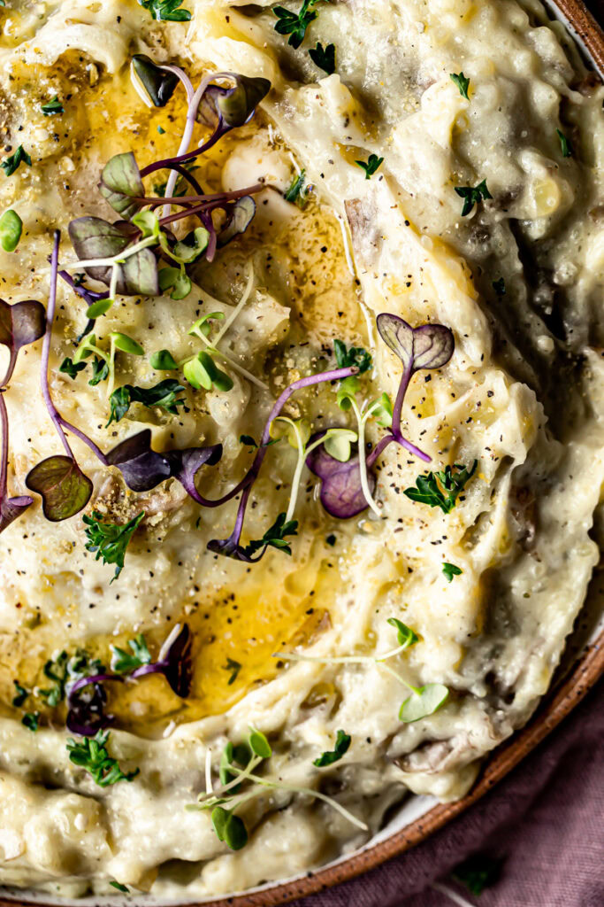 closeup of a bowl of mashed potatoes with melted butter garnished with micro greens on top of a purple linen towel