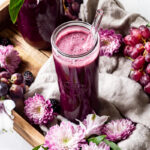 a glass of purple passion blender juice with purple flowers and grapes around it.