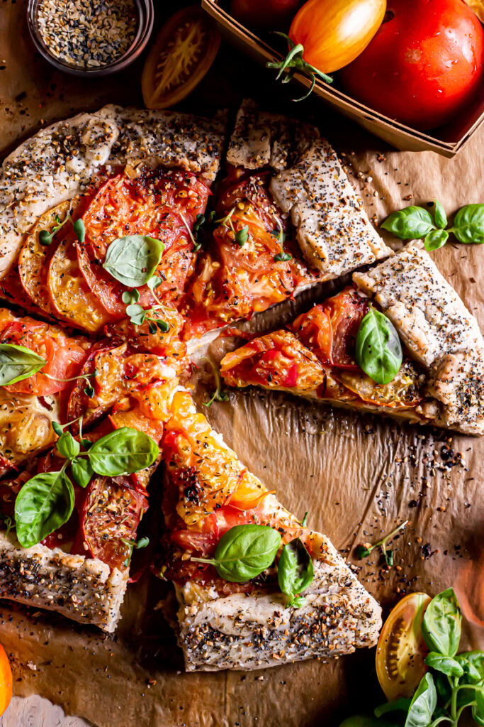 close up of a sliced tomato galette with a  basket of tomatoes beside it and fresh basil leaves