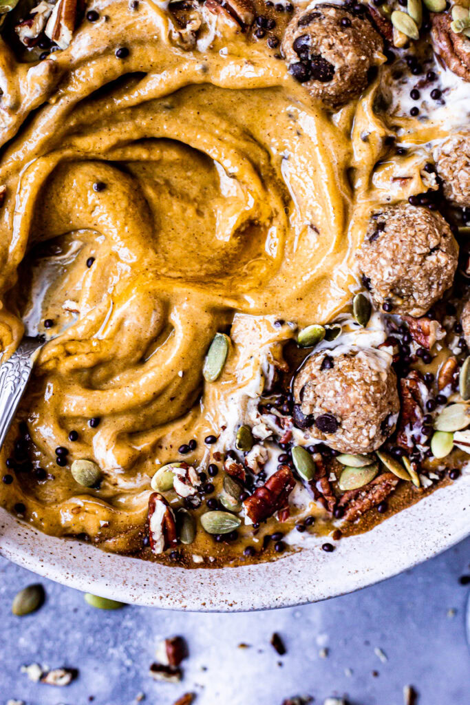 an extreme closeup of a pumpkin pie smoothie in a bowl topped with cookie dough balls, pumpkin seeds, pecans, chocolate covered chia seeds drizzled with maple syrup