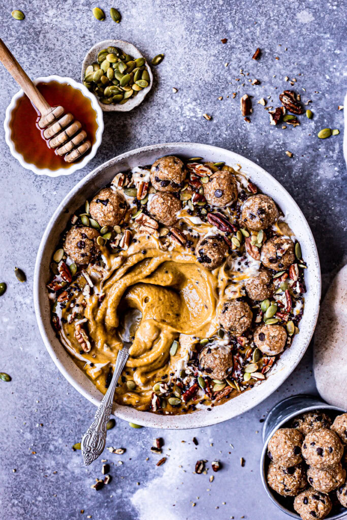 a pumpkin pie smoothie in a bowl topped with cookie dough balls, pumpkin seeds, pecans, chocolate covered chia seeds drizzled with maple syrup