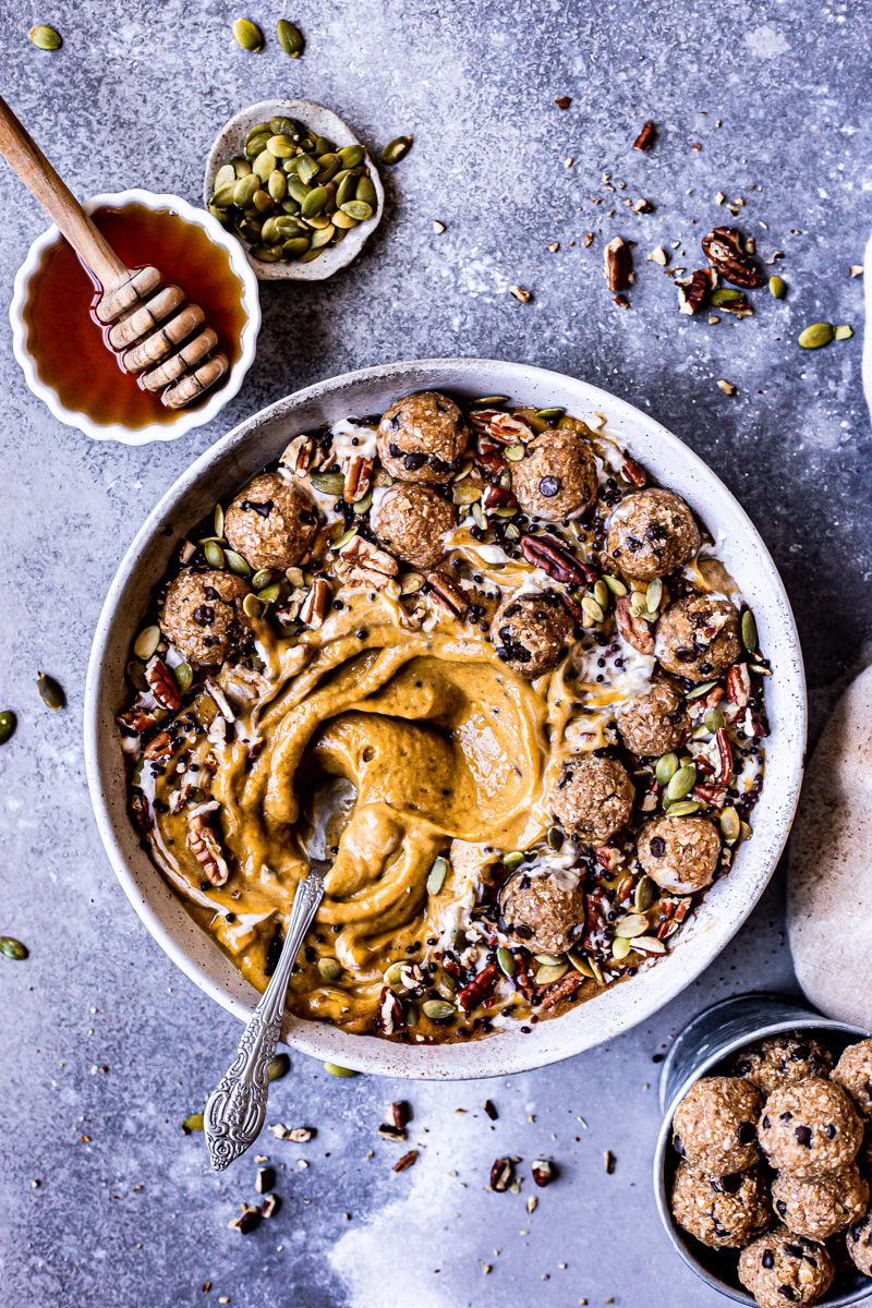 a pumpkin pie smoothie in a bowl topped with cookie dough balls, pumpkin seeds, pecans, chocolate covered chia seeds drizzled with maple syrup
