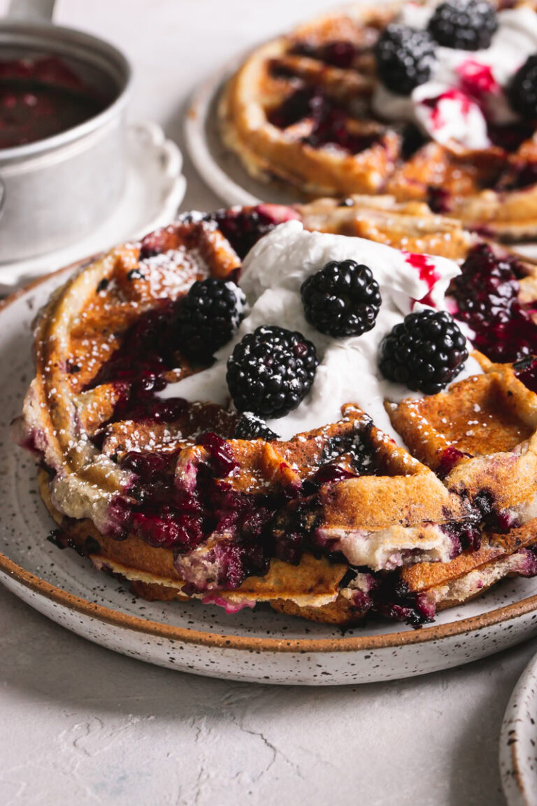 close up side view of a plate of blackberry waffles with more in the background and a pot of blackberry syrup