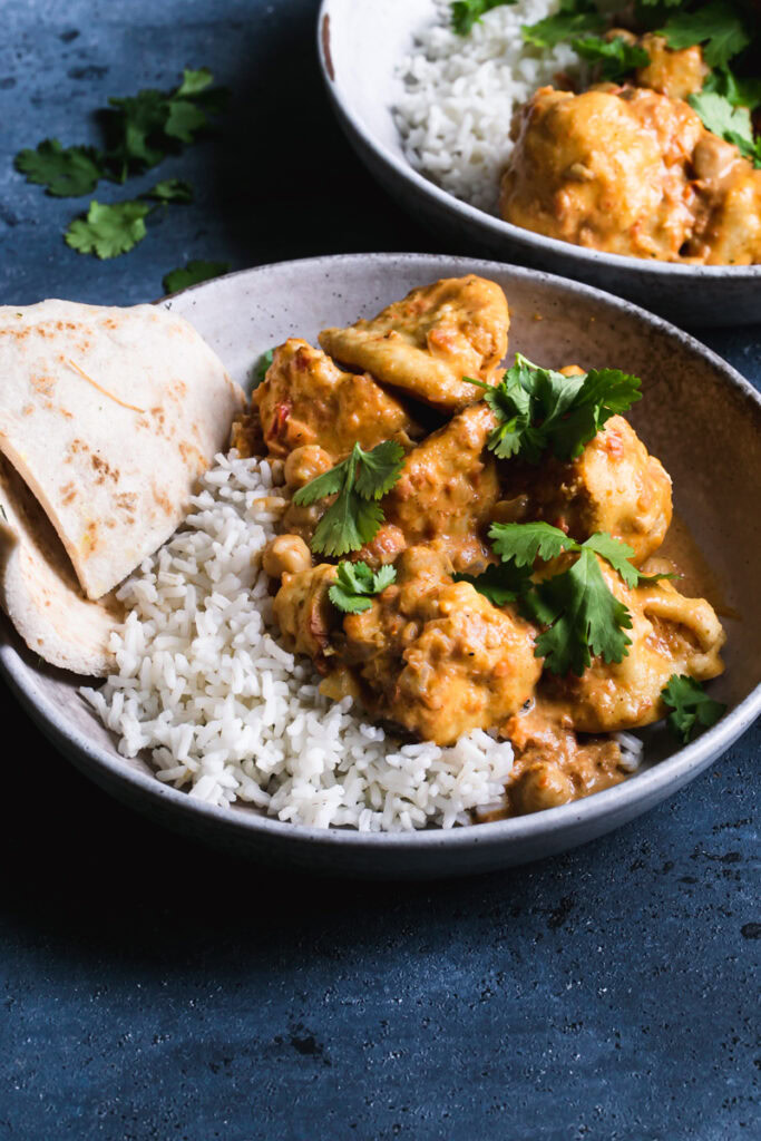 side view of two bowls of rice with breaded cauliflower tossed in a vegan Indian butter sauce garnished with fresh cilantro and a side serving of naan