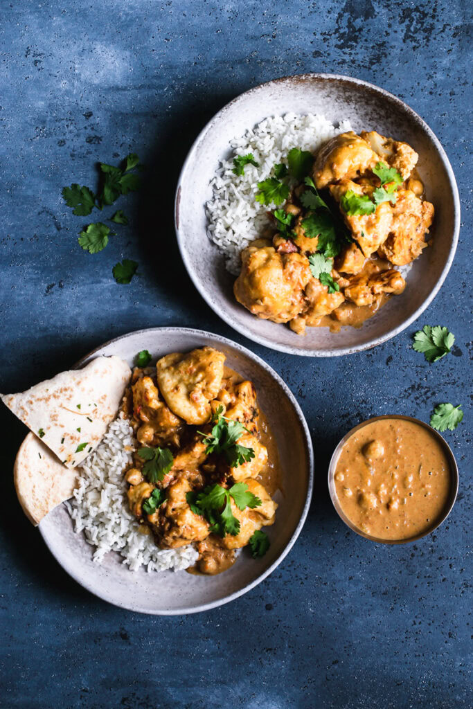 full shot of two bowls of naan butter cauliflower on top of rice, with cilantro leaves and Indian butter sauce beside it