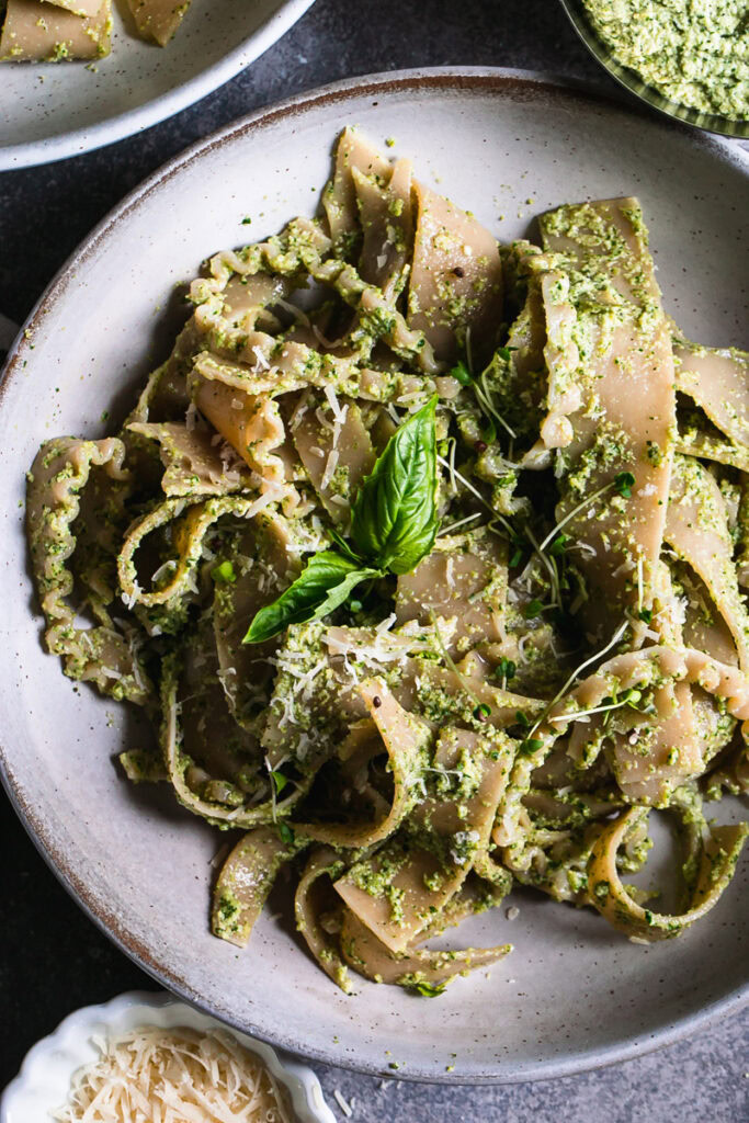 a close up of a bowl of olive pesto pasta garnished with a basil leaf a bowl of olive pesto and vegan parmesan cheese beside the bowl of pasta