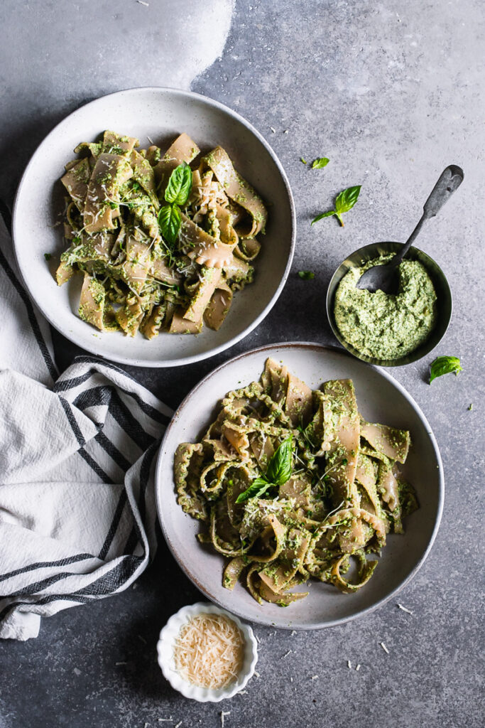 full shot of two bowls of olive pesto pasta with a linen napkin, a bowl of pesto, and vegan parmesan cheese beside it