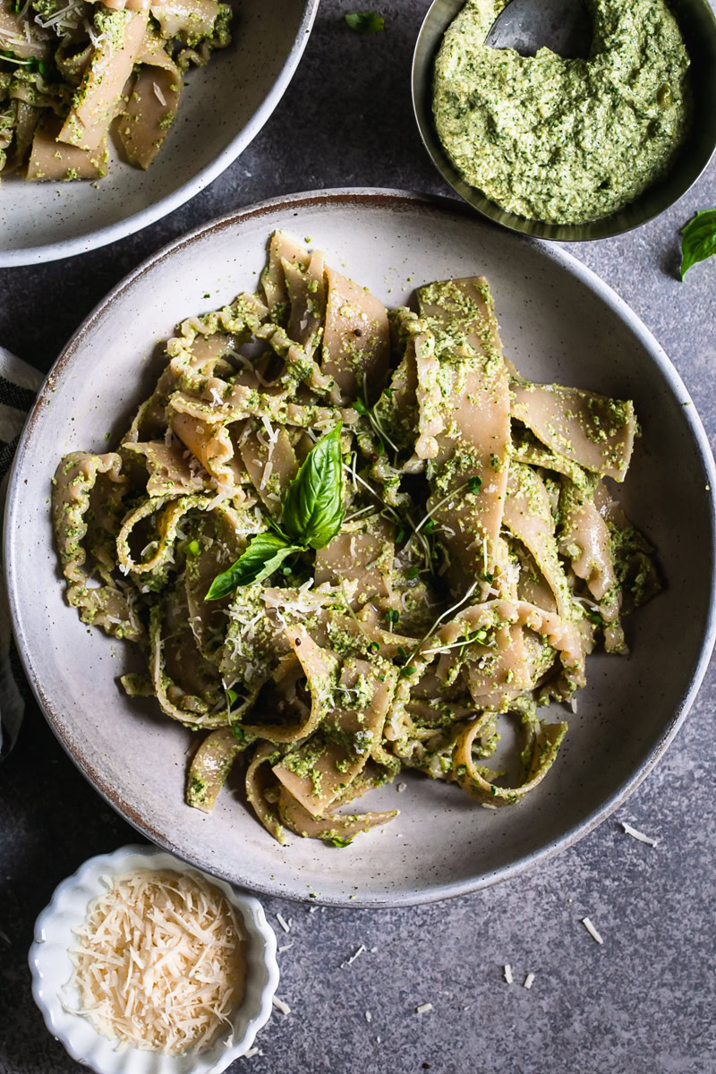 a close up of a bowl of olive pesto pasta garnished with a basil leaf a bowl of olive pesto and vegan parmesan cheese beside the bowl of pasta