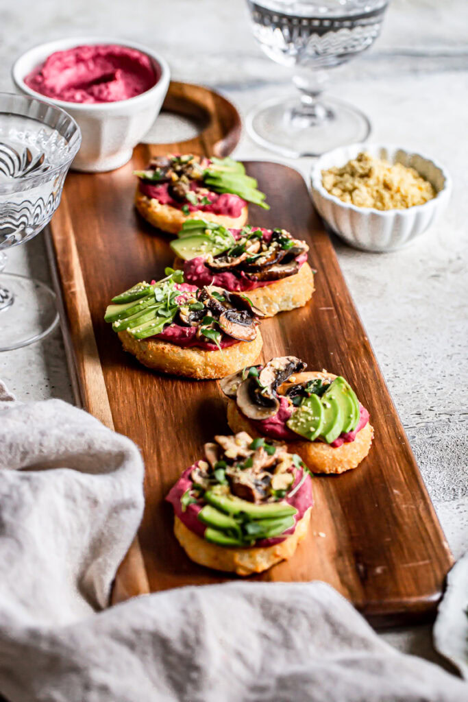 3/4 view of a wooden board with five hash browns topped with beet hummus, balsamic mushrooms, avocado, vegan parmesan cheese, and micro greens with a bowl of beet hummus and water next to it