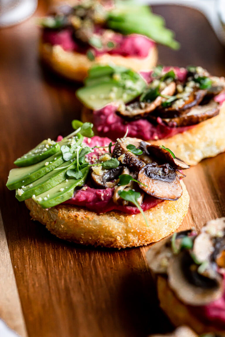 close up of hash browns topped with beet hummus, sliced avocado, balsamic roasted mushrooms, vegan parmesan cheese and micro greens