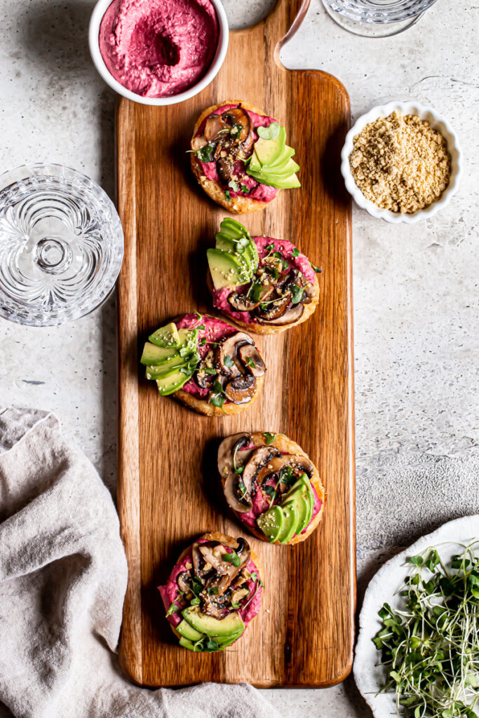 a wooden board with five hash browns topped with beet hummus, balsamic mushrooms, avocado, vegan parmesan cheese, and micro greens with a bowl of beet hummus and water next to it
