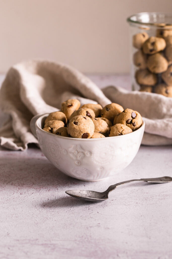 a mug full of baked cookie dough balls with a spoon in front, a lined napkin behind and a jar of leftover cookie dough balls in the background.