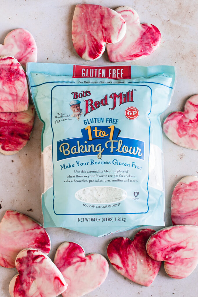 11 heart shaped sugar cookies with red, pink and white marble icing and a bag of bob's red mill gluten-free 1 to 1 baking flour in the center of the cookies