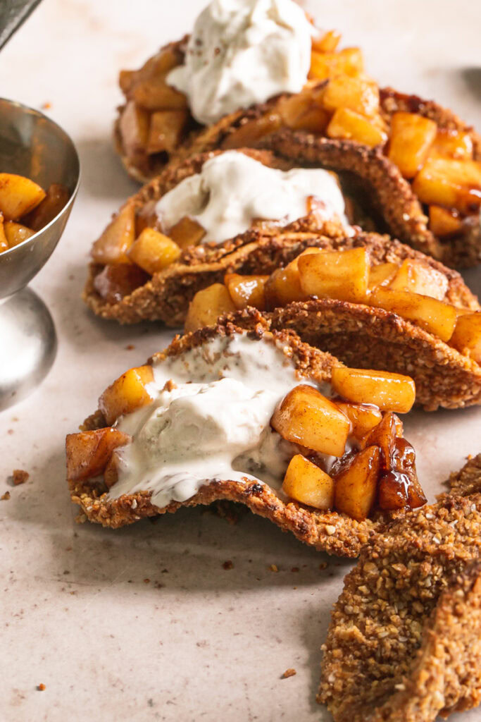 3/4 image of apple pie tacos with partially melted ice cream on top and a bowl of apple pie filling in the background