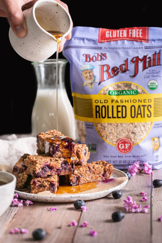 head on view of Blueberry Oatmeal French Toast Sticks piled on top of one another pouring maple syrup over them with a bag of Bob's Red Mill Organic Old Fashioned Rolled Oats in the background