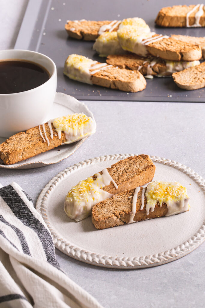 two lemon vanilla biscotti on a plate with a cup of coffee and another biscotti behind it and a cookie sheet of biscotti in the background