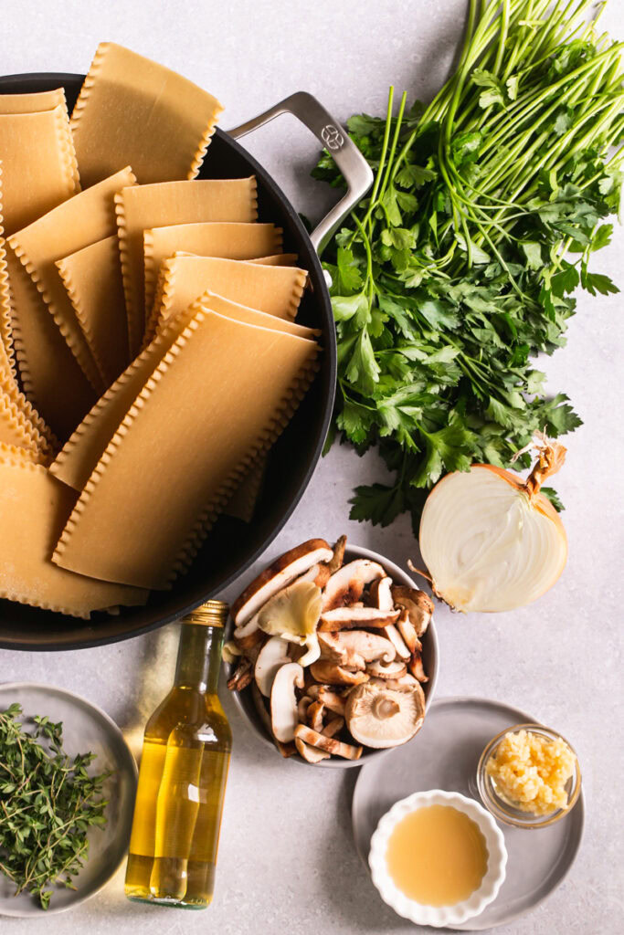 a stock pot with lasagna uncooked lasagna noodles, next to it fresh parsley, thyme, a bowl of lemon juice and minced garlic, a bowl of mushrooms and a bottle of white wine
