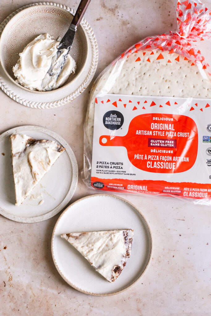 two slices of cinnamon roll pizza with a bowl of vegan cream cheese icing in the upper left and the package of little northern bakehouse pizza crusts in the upper right