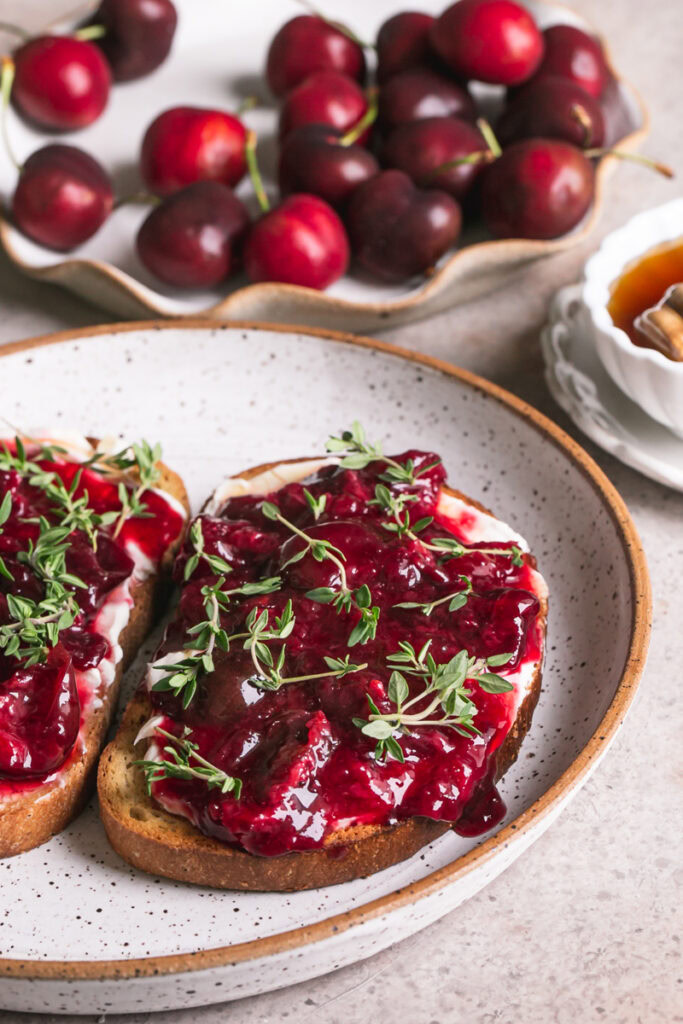 a close up 3/4 shot of two slices of toast with dairy-free cream cheese, roasted cherries, fresh thyme on a plate with a plate of fresh cherries in the background