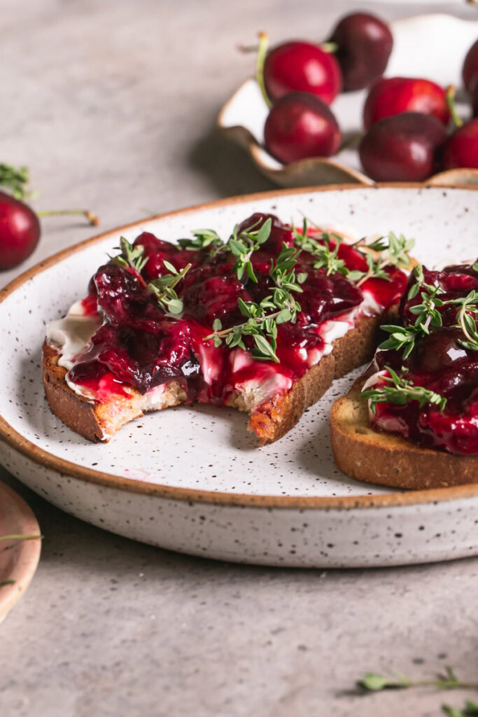 a close up 3/4 shot of two slices of toast with dairy-free cream cheese, roasted cherries, fresh thyme on a plate one slice with a bite taken out of it and a plate of fresh cherries in the background