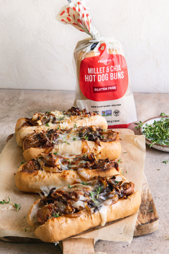 side view of vegan French onion brats on a parchment lined wooden cutting board with a bag of little northern bakehouse hot dog buns in the background