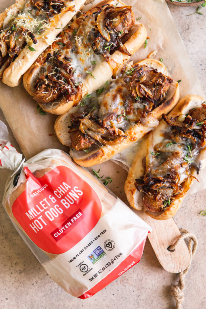 four vegan French onion brats layed out on a parchment line wooden cutting board and a bag of little northern bakehouse hot dog buns next to it
