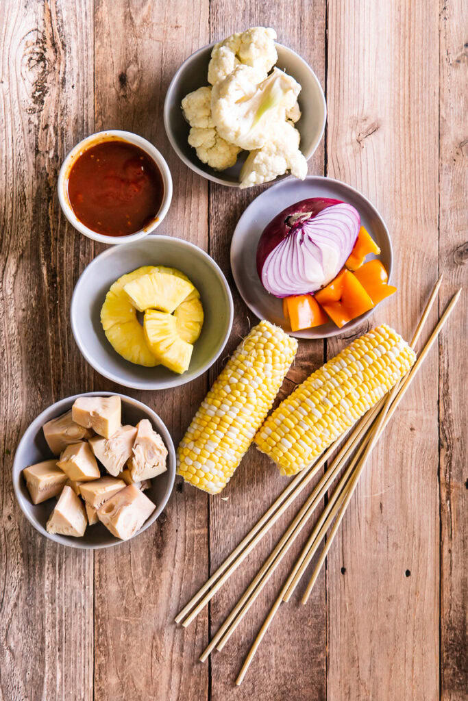 kabob ingredients all laid out- corn on the cob, jackfruit, pineapple, red onion, cauliflower, pepper, kabob sticks and homemade barbecue sauce