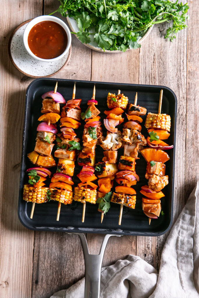 Grilled veggie barbecue jackfruit kabobs in a square Calphalon grilling pan with a bowl of barbecue sauce beside it and a plate of cilantro above it