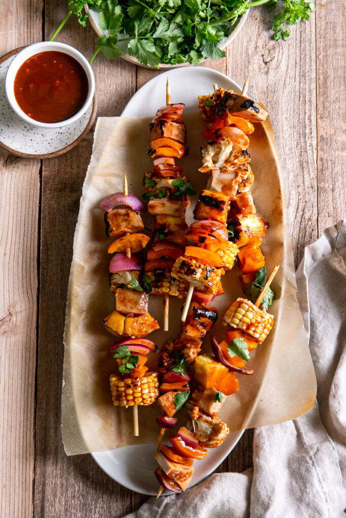 an oval platter with grilled veggie barbecue kabobs on it with a side of barbecue sauce in a bowl beside it and fresh cilantro on a plate above it