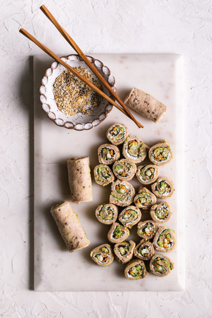 a marble cutting board with 3 rolled up unsliced veggie sushi sandwiches and sliced rollup sandwiches with a bowl of everything bagel spice and wooden chopsticks