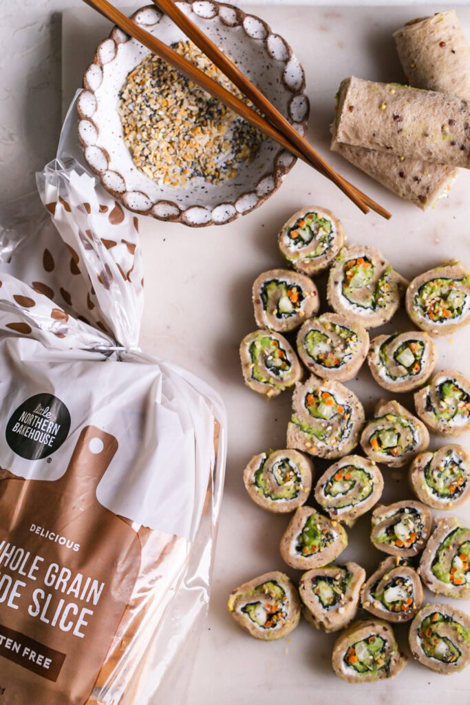 a marble cutting board with 3 rolled up unsliced veggie sushi sandwiches and sliced rollup sandwiches with a bowl of everything bagel spice and wooden chopsticks an a loaf of Little Northern Bakehouse whole grain bread