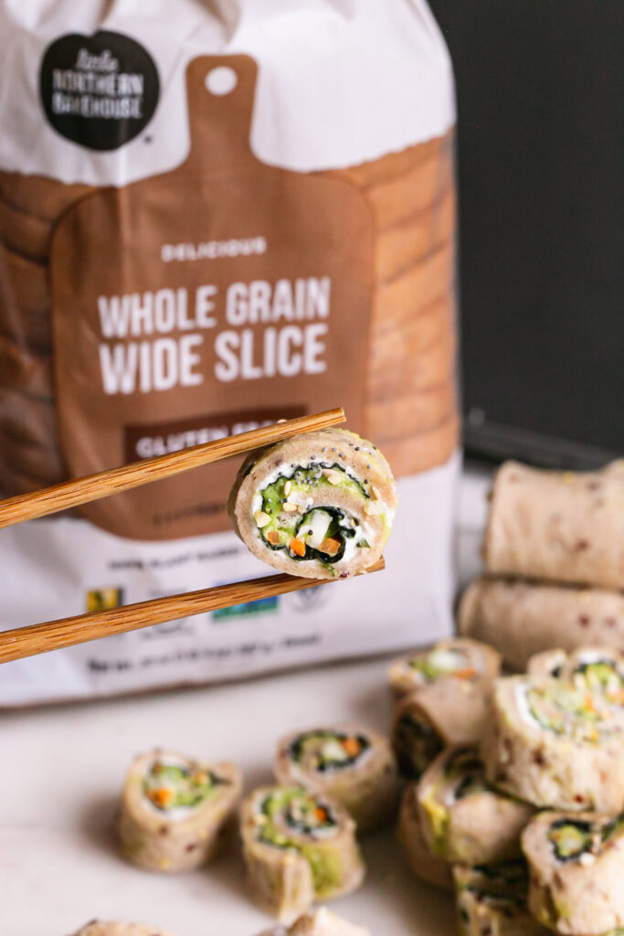 vertical image of a slice of veggie sandwich sushi held with chopsticks and a loaf of Little Northern Bakehouse whole grain bread in the background
