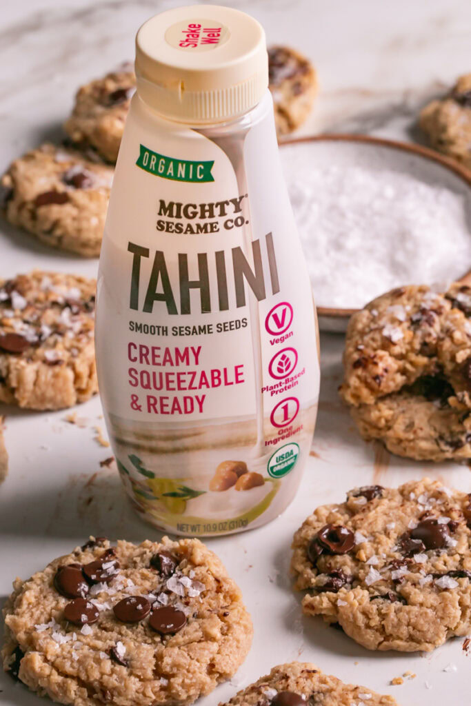 3/4 view of a bottle of mighty sesame co organic tahini with flourless salted tahini chocolate chip cookies surrounding it and a plate of salt