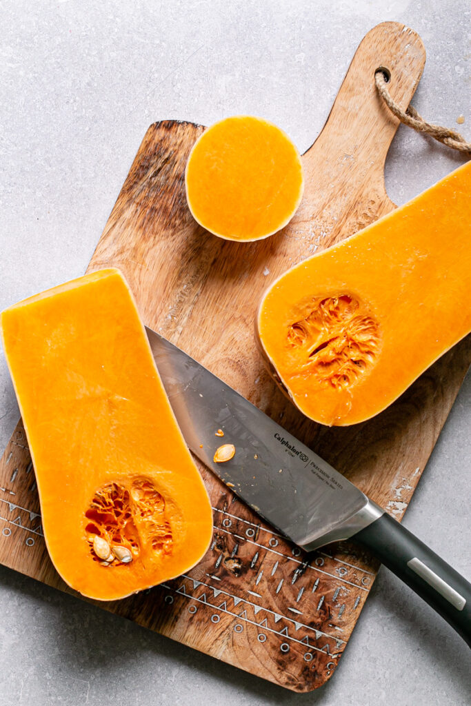a butternut squash cut in half on a cutting board with the knife laying beside it