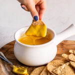 a bowl of vegan nacho cheese sauce with a chip being dipped in it and cheese sauce dripping off of the chip