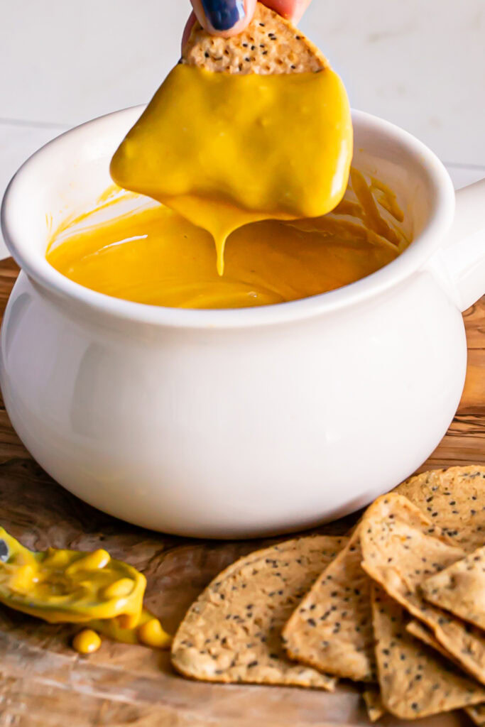 a close up of a bowl of vegan nacho cheese sauce with a chip being dipped in it and cheese sauce dripping off of the chip