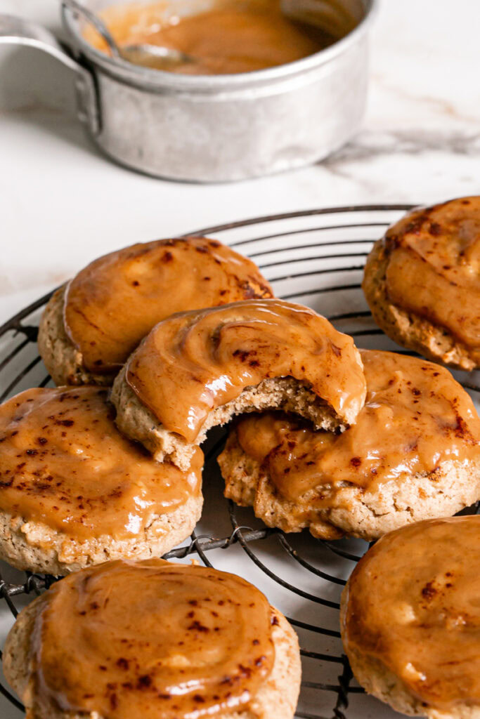 3/4 view of caramel apple cider cookies, one with a bite taken out of it and a saucepan of caramel in the background