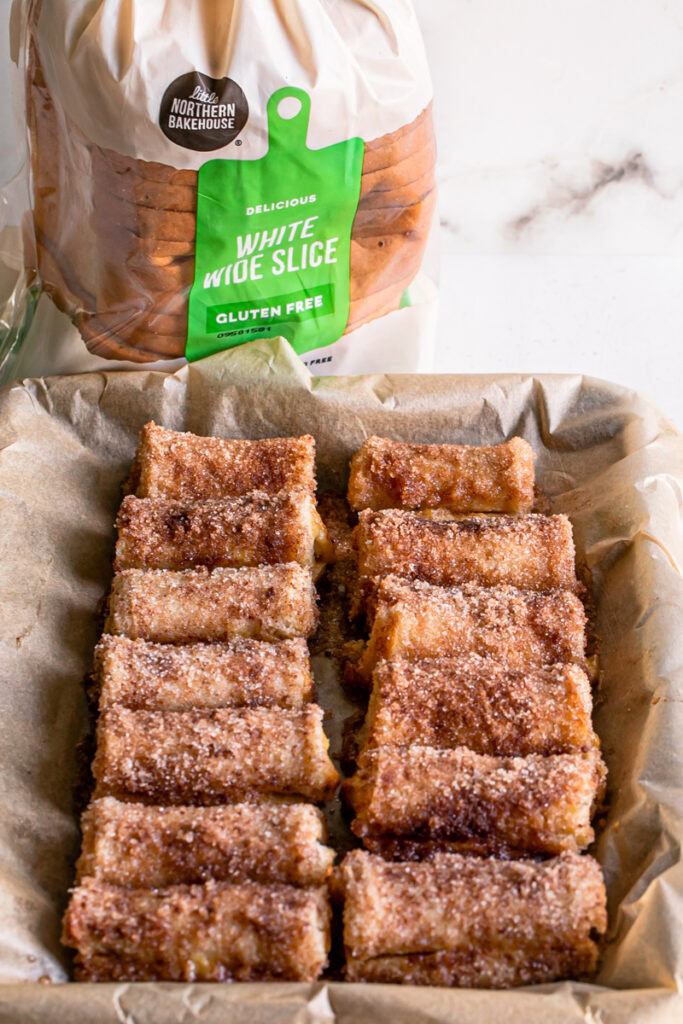 Pumpkin Cream Cheese Rollups lined up in a baking tray lined with parchment paper and a loaf of little northern bakehouse white wide sliced gluten-free bread in the background