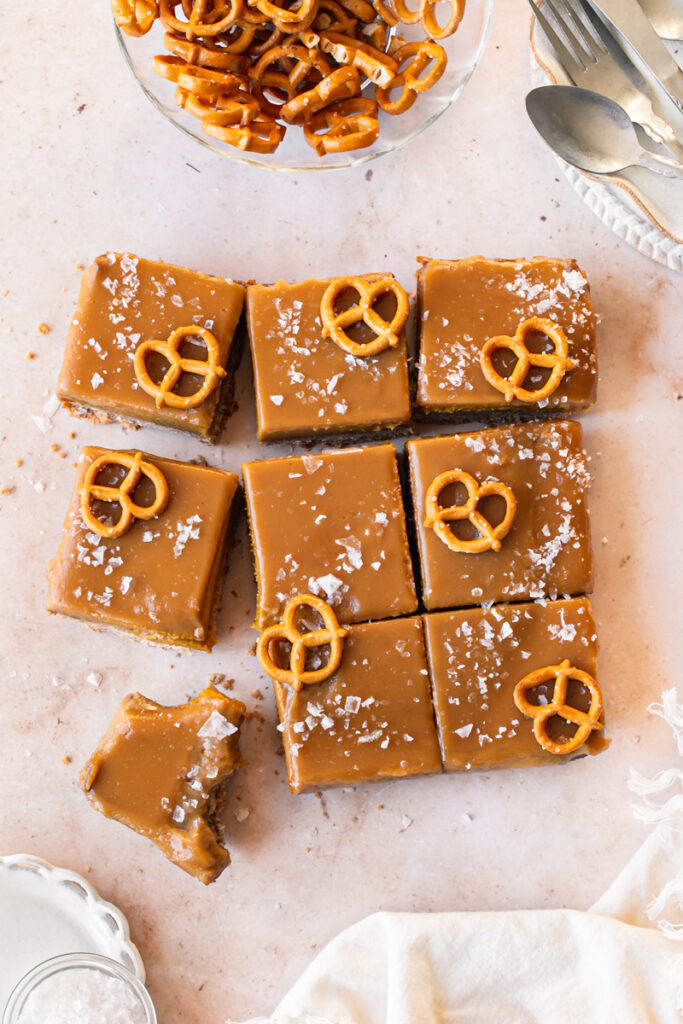 flatlay shot of slices of salted pumpkin pie bars with pretzels on top a bowl of pretzels, plates, a bowl of salt and a napkin