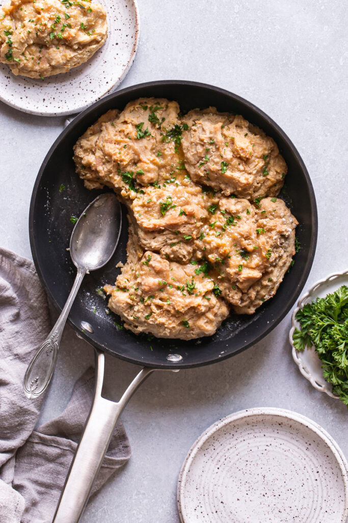 Vegan Gluten-Free Buttermilk Skillet Biscuits in the skillet with plates, a plate of fresh parsley, and one biscuit served on a plate