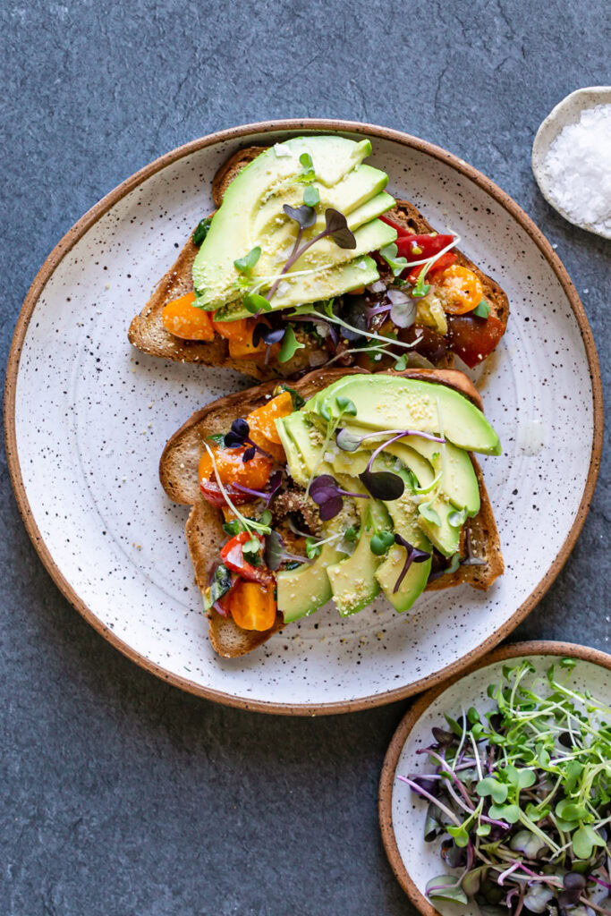 two slices of Bruschetta Avocado Toast on a plate with a plate of micro greens next to it and a pinch plate of flake salt above it