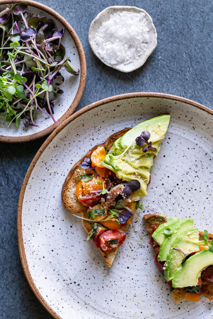 a slice of 3/4 view of two slices of Bruschetta Avocado Toast on a plate sliced in half diagonally with a plate of micro greens and a pinch plate of flake salt above it